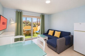 Ein kompakter Wohnraum mit einer Couch, einem kleinen Tisch, einem Glastisch, einem weißen Kühlschrank und Blick durch eine Glastür zu einem Außenbereich mit Tisch und Stühlen.