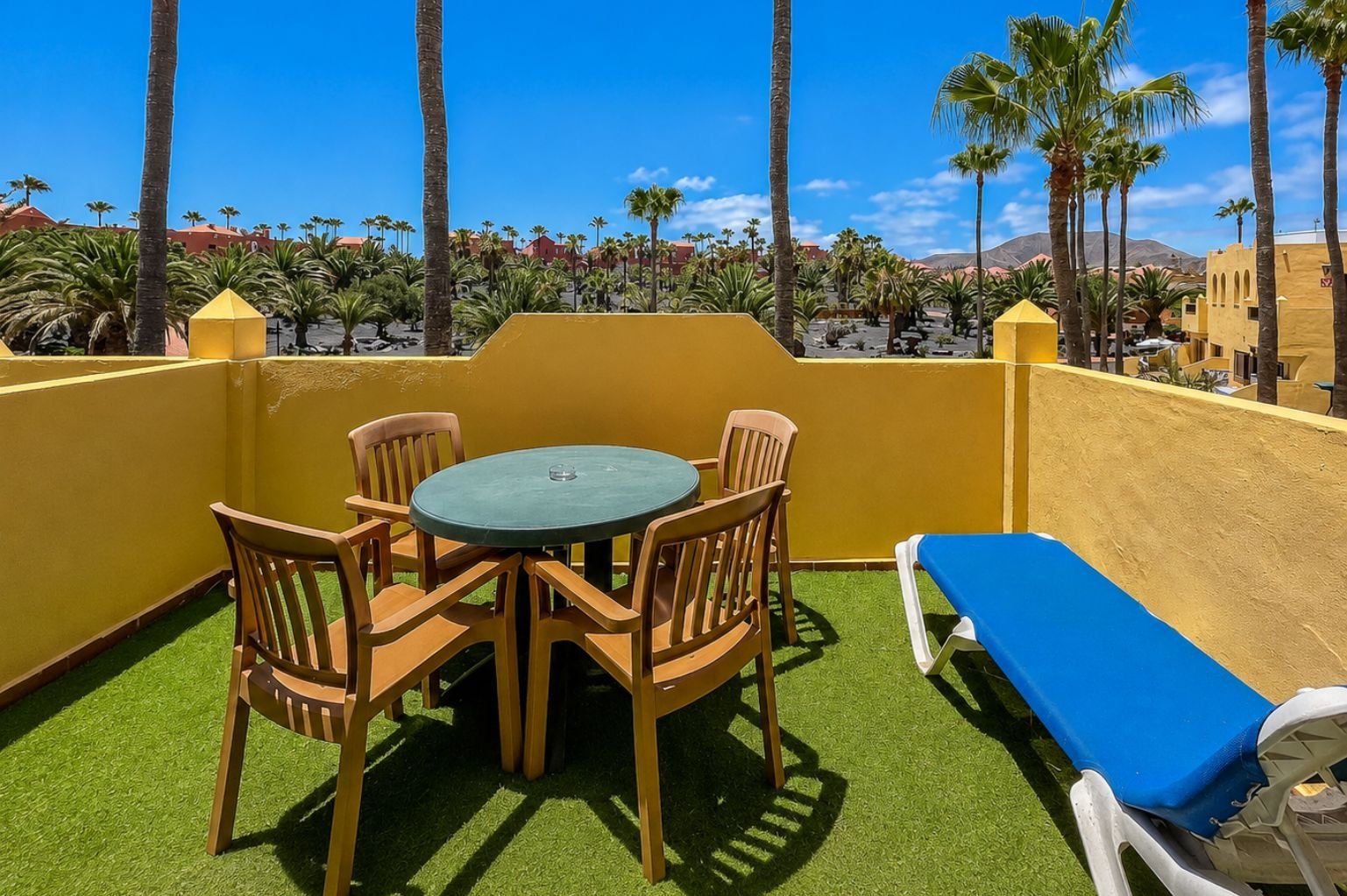 A balcony with a round table and chairs, a blue lounge chair, and a view of palm trees and buildings.