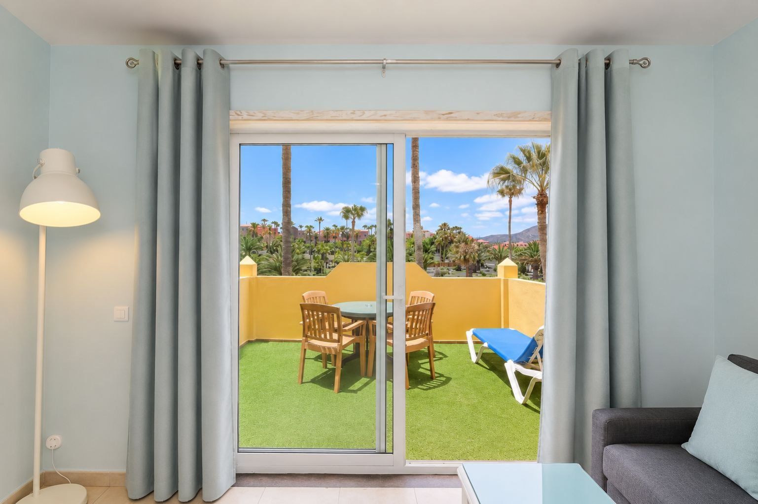 Quarto com porta de vidro deslizante que dá para área externa com mesa, cadeiras, sofá e luminária.
