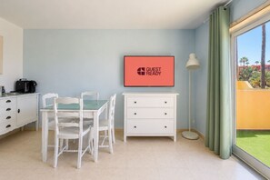 Ein Zimmer mit Esstisch, Stühlen, einer weißen Kommode, einem gerahmten Schild, einer Stehlampe und Blick auf Palmen durch ein Fenster.