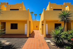 A courtyard with yellow buildings, a tiled walkway, and some greenery. - GuestReady - Modern Stay with Pool and Terrace (Corralejo)