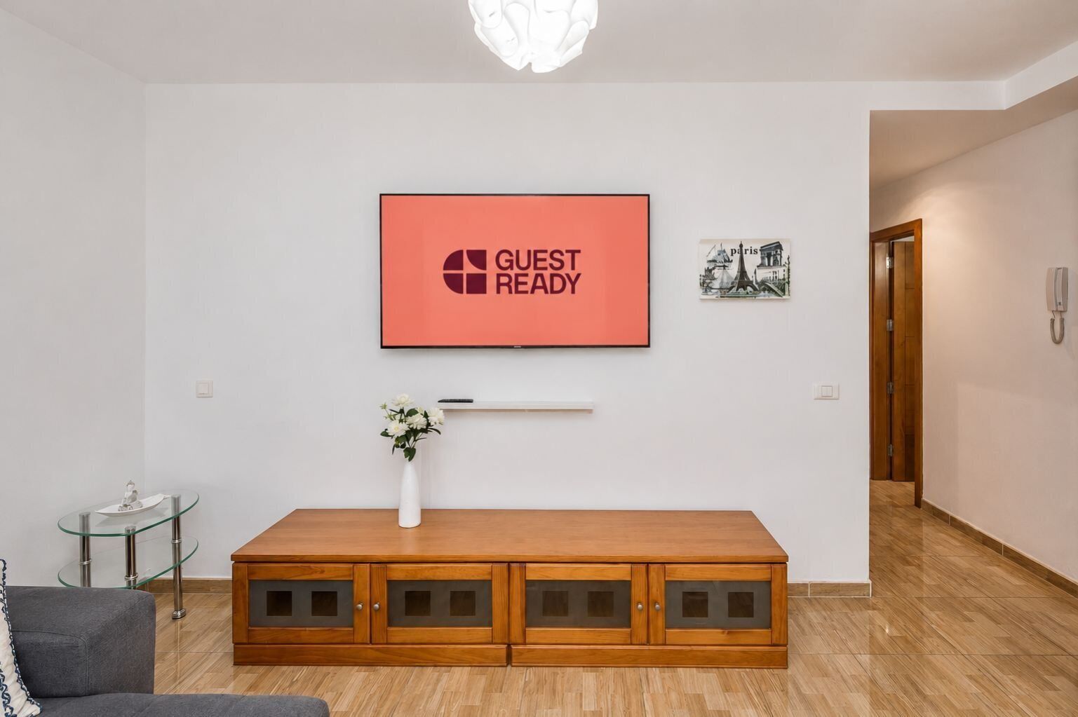 Ein modernes Wohnzimmer mit einem Holzkabinett, einem Glastisch, einer Blumenvase, einem gerahmten Bild und einem Schild mit der Aufschrift 'GUEST READY' an der Wand.