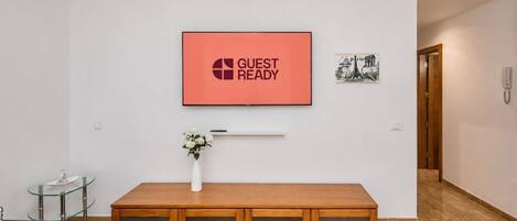 A modern living room with a wooden cabinet, a glass table, a vase with flowers, a framed picture, and a 'GUEST READY' sign on the wall.