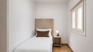 Una habitación pequeña y estrecha con una cama individual, una mesita de noche con lámpara y una ventana.