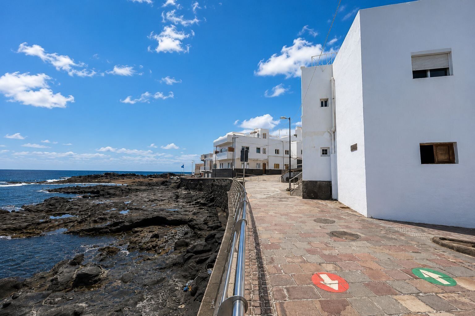 A coastal walkway with a metal railing, leading to white buildings with multiple windows, and a clear blue sky.