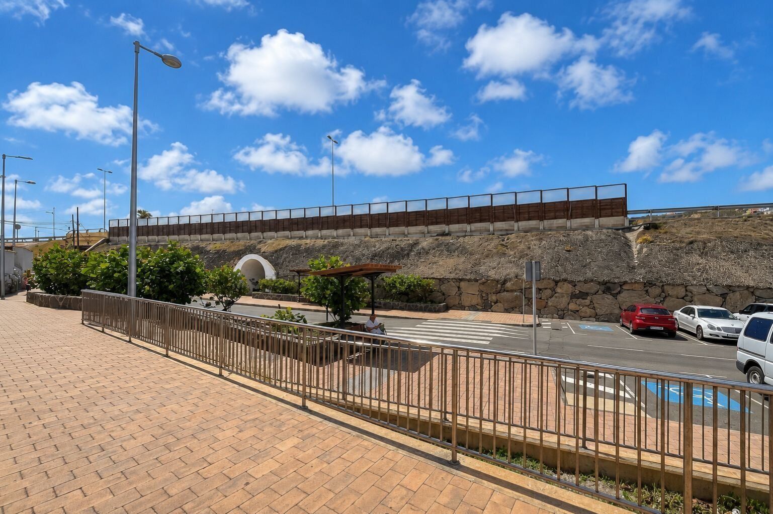 A paved walkway with a metal fence, a covered seating area, and a stone wall in the background.