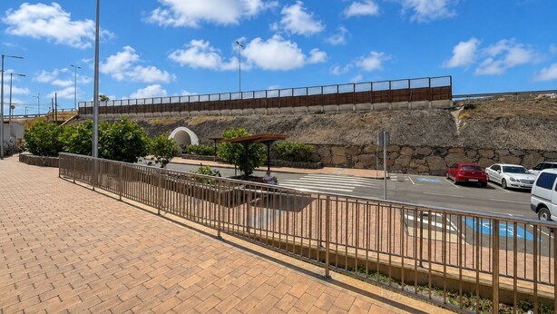 Un sendero pavimentado con una cerca metálica, una zona de estar techada y un muro de piedra al fondo.