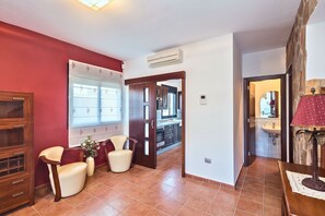 A hotel room with a red wall, a wooden cabinet, a seating area with two chairs, a small table with a vase, and a view into the kitchen through an open door. - GuestReady - Villa Datana with Pool (Mogán)