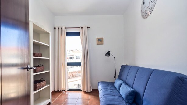 Een kamer met een blauwe bank, een kledingkast met planken, een deur, een raam met gordijnen, en een wandklok.