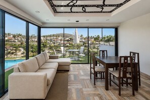 A modern living room with a large window, a sectional sofa, a dining table with chairs, and a view of a residential area. - GuestReady - Villa Datana with Pool (Mogán)