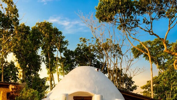 Un edificio de forma abovedada con una puerta de madera, rodeado por un jardín y una piscina circular con borde de madera.