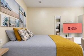 Une chambre d’hôtel avec un lit, un téléviseur affichant le message « CHAMBRE PRÊTE POUR L’ARRIVÉE DES CLIENTS », et un mur orné d’œuvres d’art encadrées.