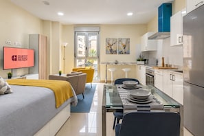 Quarto de hotel moderno com cama, mesa de jantar para dois, área de cozinha e televisão exibindo 'GUEST READY'.