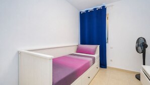 Yhden hengen huone, jossa on valkoinen pääty, yöpöytä, tuuletin ja sininen verho.