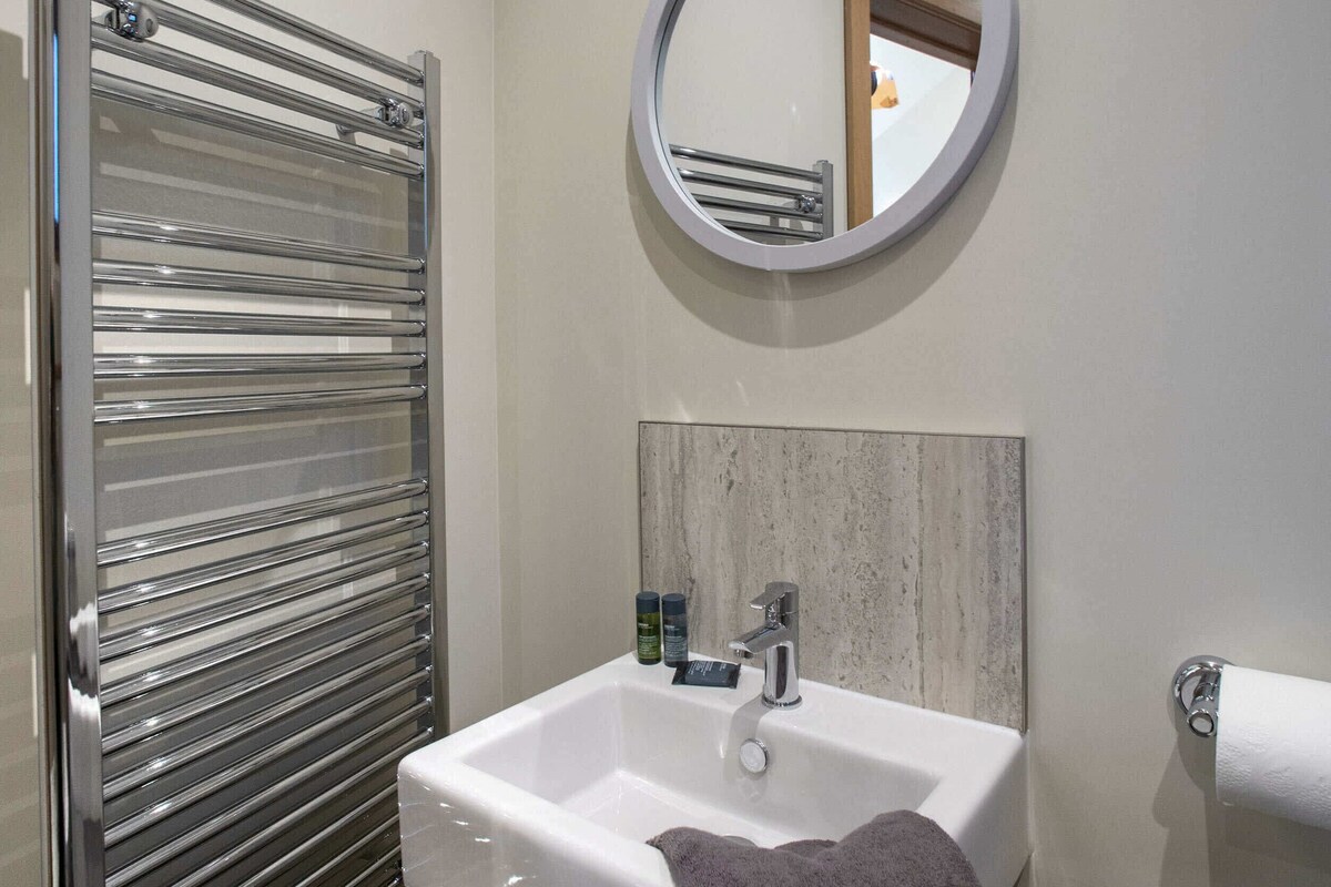 A bathroom with a glass shower enclosure, a white sink with a wooden panel above it, and a round mirror on the wall.