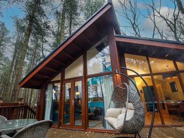 Una cabaña moderna con puerta de cristal, una silla colgante y una terraza de madera.