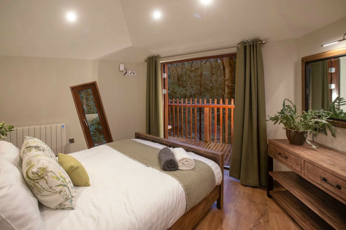 A bedroom with a bed, a wooden dresser, a mirror, and a window with a view of trees.