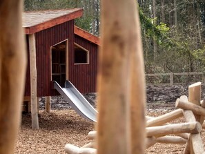 Una casita de juegos de madera con un tobogán, rodeada por una valla de madera y acolchado de mantillo.