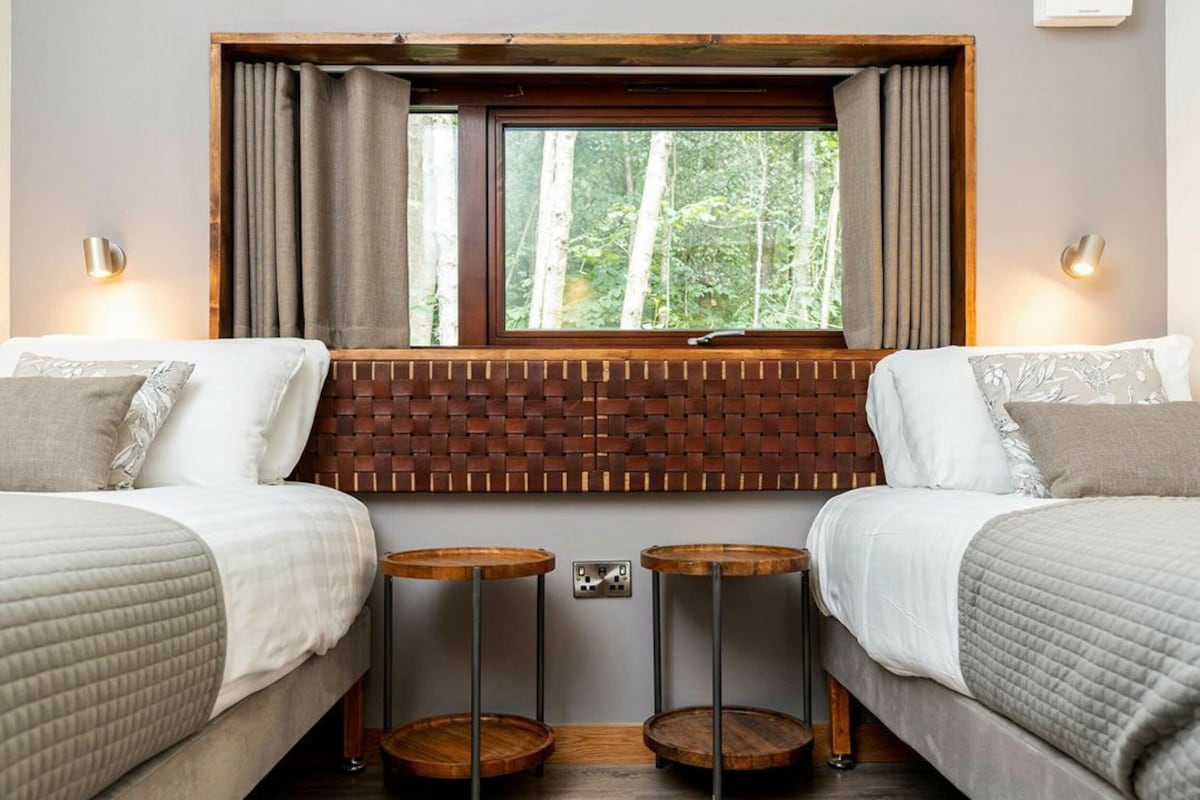 A bedroom with two beds, a window with curtains, a brick-patterned headboard, and two small wooden tables.