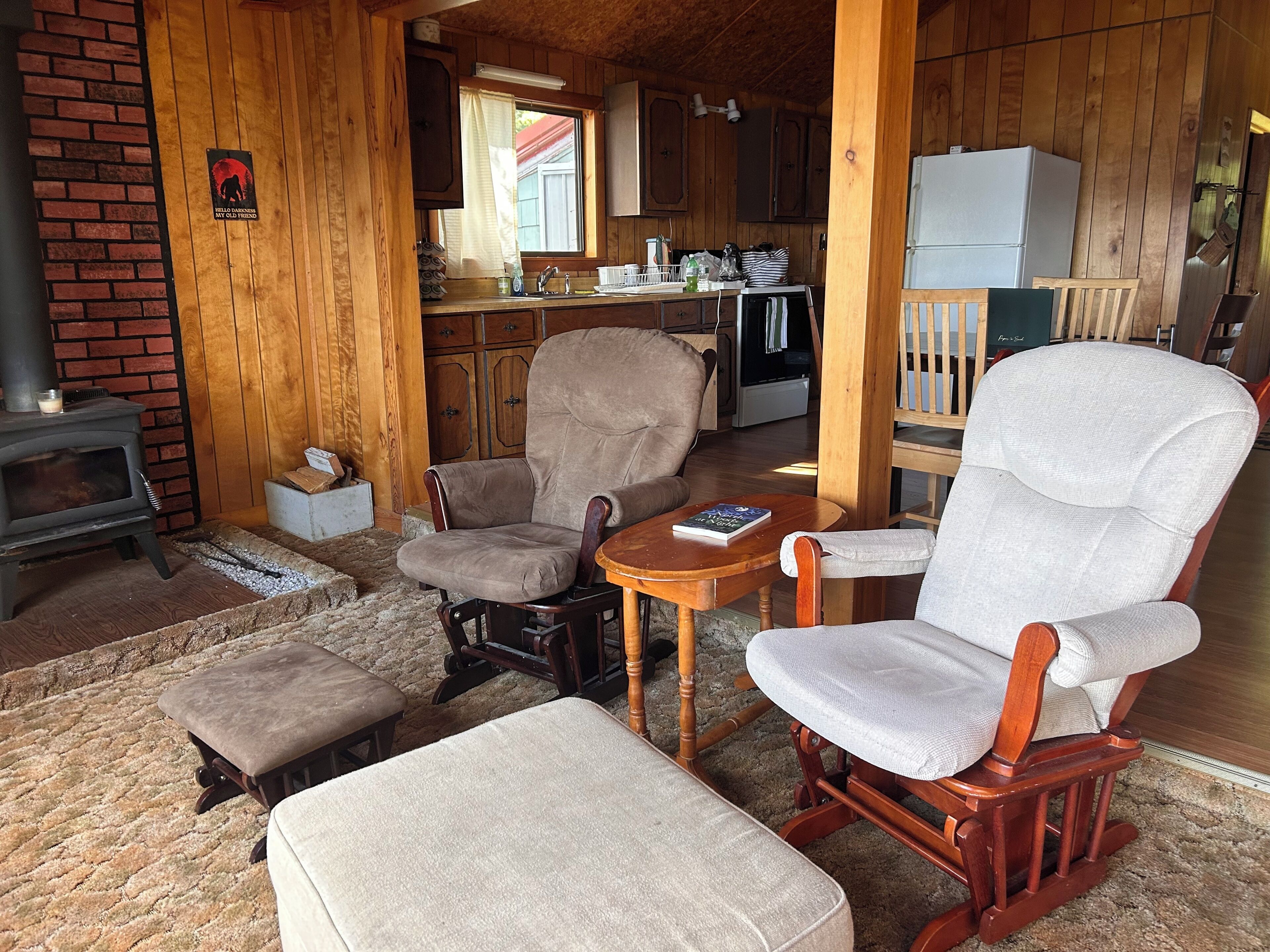 A cozy cabin interior with a fireplace, wooden furniture, a kitchen area, and a window with curtains.