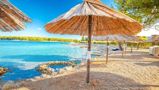 A beach with thatched umbrellas, clear blue water, and a rocky shoreline. - Sun-Kissed Sailing Adventure in Northern Croatia (Punat)