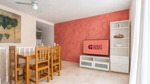 Una habitación con una mesa de comedor y sillas, un televisor sobre un mueble bajo y un estante blanco.