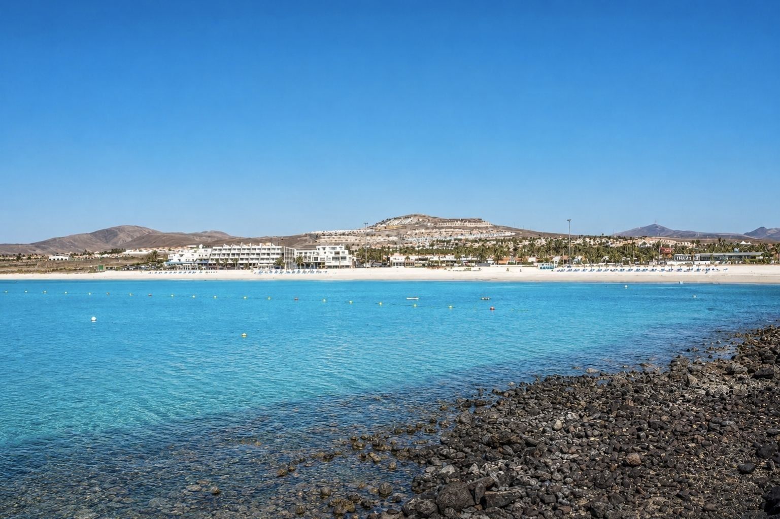 Een hotel aan de kust met uitzicht op het strand, een rotsachtige kustlijn en een berg op de achtergrond.
