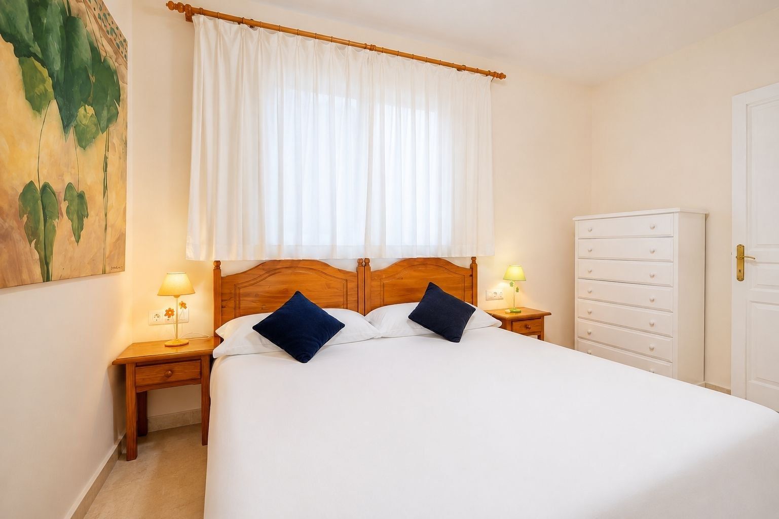 Une chambre d’hôtel avec un grand lit, deux tables de chevet avec des lampes, une tête de lit en bois, une armoire blanche et un tableau au mur.