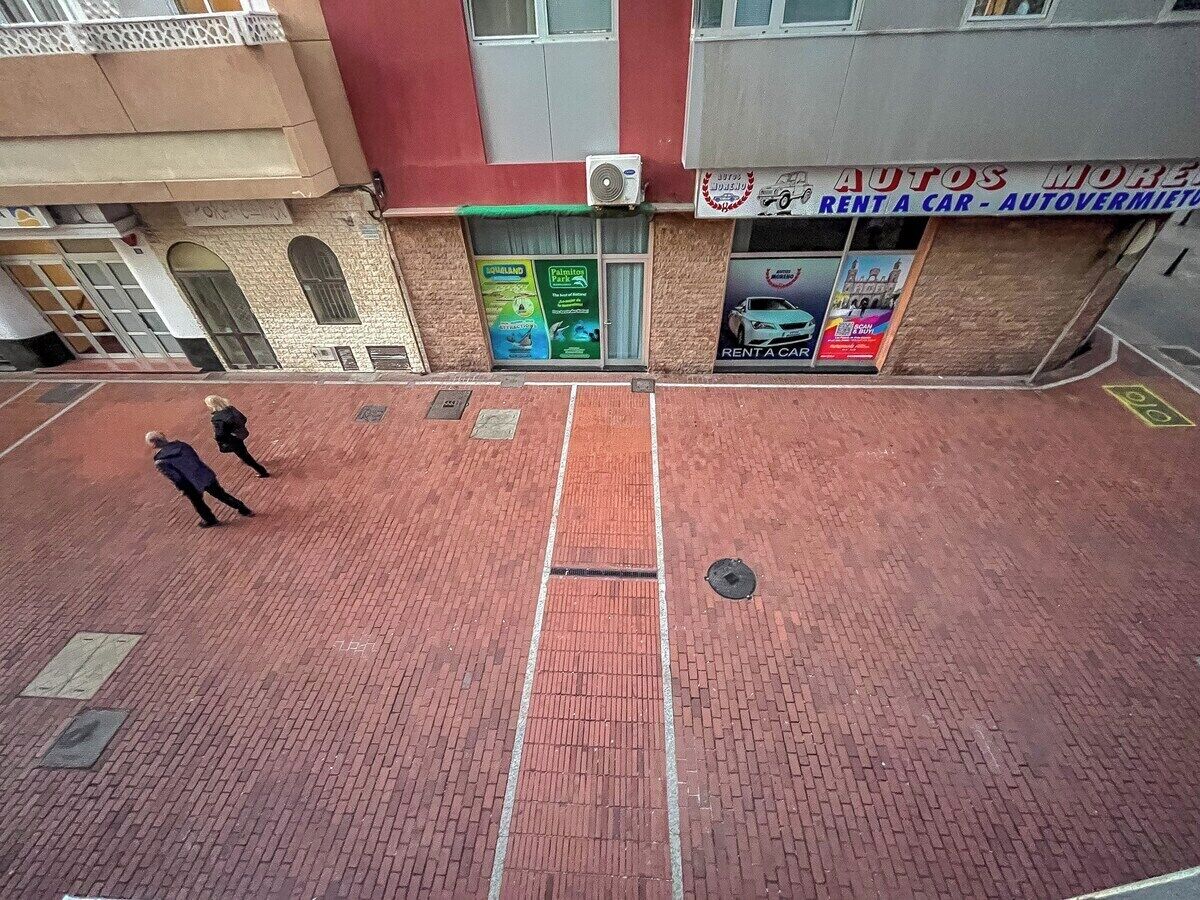 A red brick pavement with a manhole cover, a building with a sign for 'Autos More' and 'Rent a Car - Autovermietung', and a green sign for 'Aquiland Park'.
