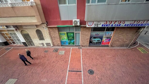 A red brick pavement with a manhole cover, a building with a sign for 'Autos More' and 'Rent a Car - Autovermietung', and a green sign for 'Aquiland Park'. - GuestReady - Salvador Cuyas Beach Walk (Las Palmas de Gran Canaria)