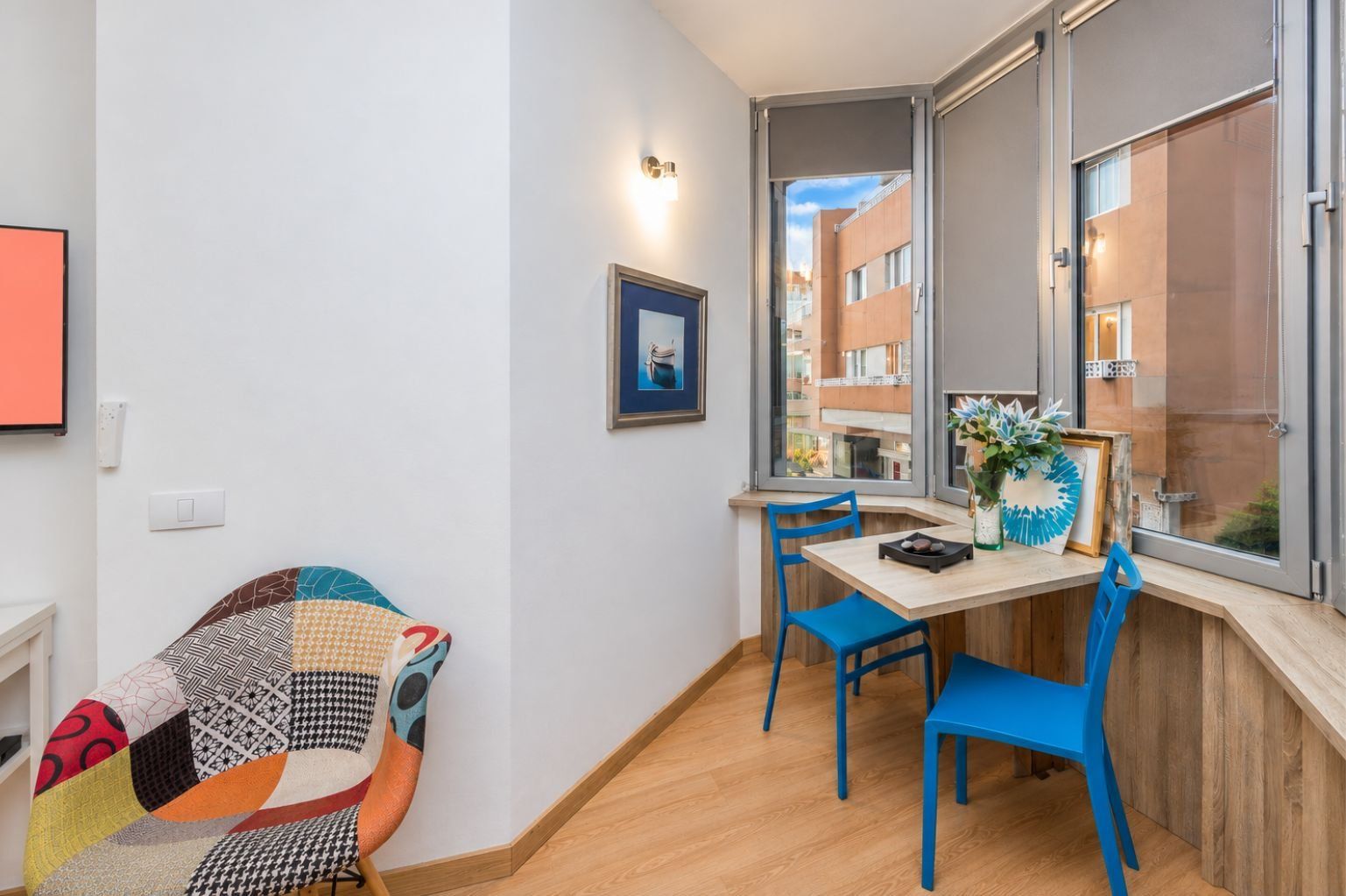 A room with a wooden floor, a table with chairs, a framed picture on the wall, and a window with a view of buildings.