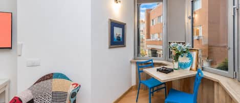 Une pièce avec un plancher en bois, une table entourée de chaises, un tableau encadré accroché au mur et une fenêtre donnant sur des immeubles.