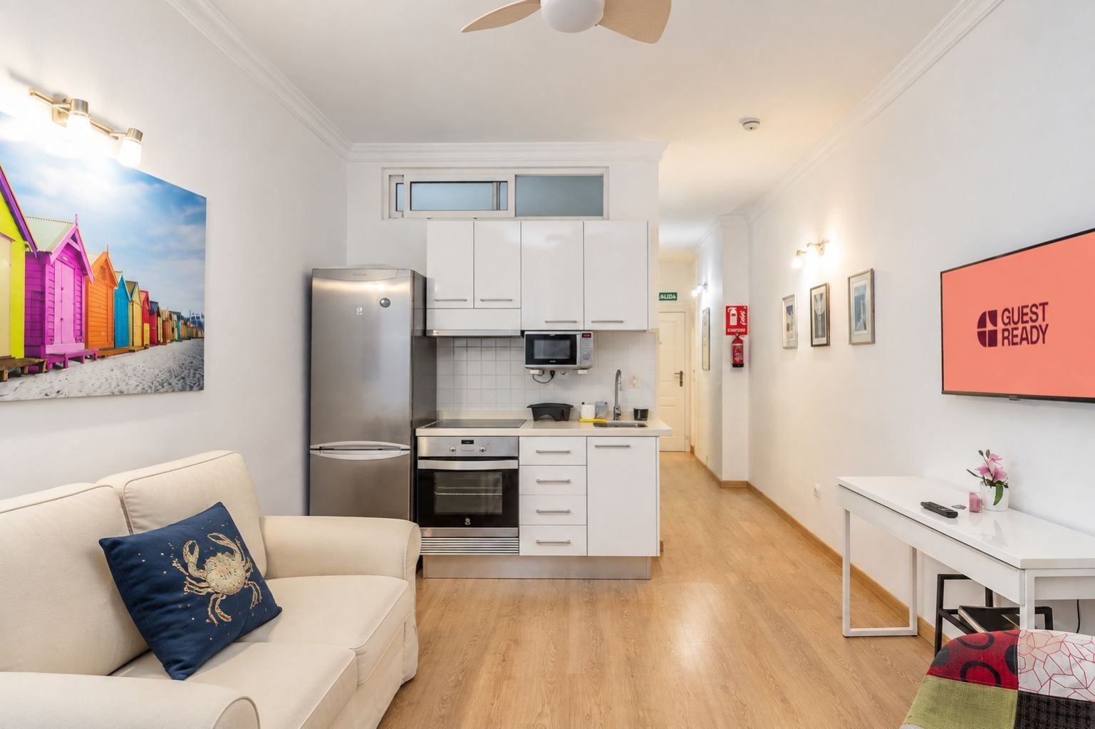 Una habitación de hotel moderna con kitchenette, un sofá con cojín decorativo, un televisor de pantalla plana y un cartel que dice 'GUEST READY'.