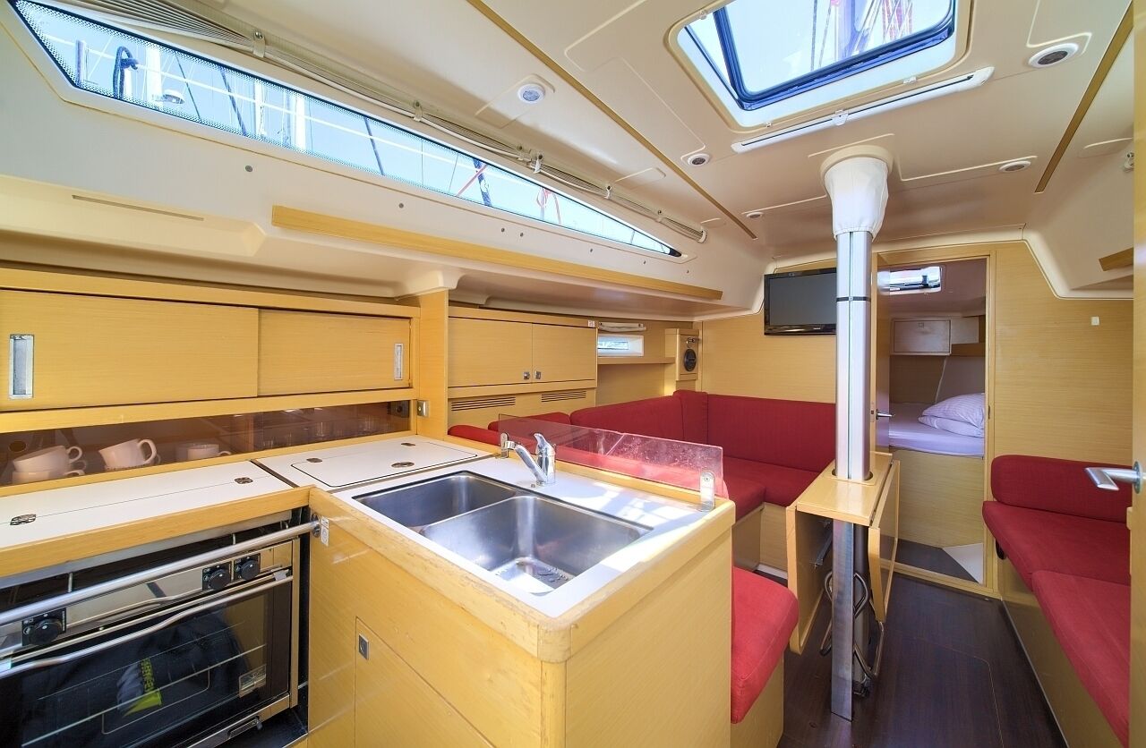 Een kleine keuken met een spoelbak, fornuis en bovenkastjes, een zithoek met rode kussens, en een televisie aan de muur.