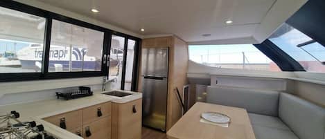 Een compacte keuken met een spoelbak, fornuis en koelkast, een eethoek met een tafel en bank, en grote ramen met uitzicht op de omgeving.