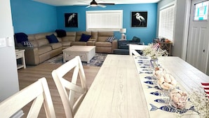 Una sala de estar con un sofá modular, una mesa de comedor con mantel decorativo, un ventilador de techo y obras de arte colgadas en la pared.
