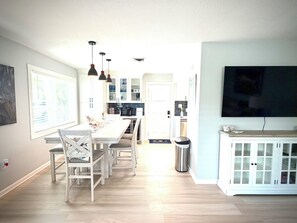 Eine moderne Küche mit weißen Schränken, eine Essecke mit einem weißen Tisch und Stühlen, ein an der Wand befestigter Fernseher und ein Mülleimer.