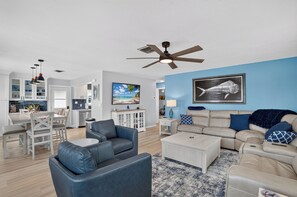 Ein modernes Wohnzimmer mit Deckenventilator, Essbereich und einer Küche im Hintergrund, sowie ein großes Fischbild an der Wand.