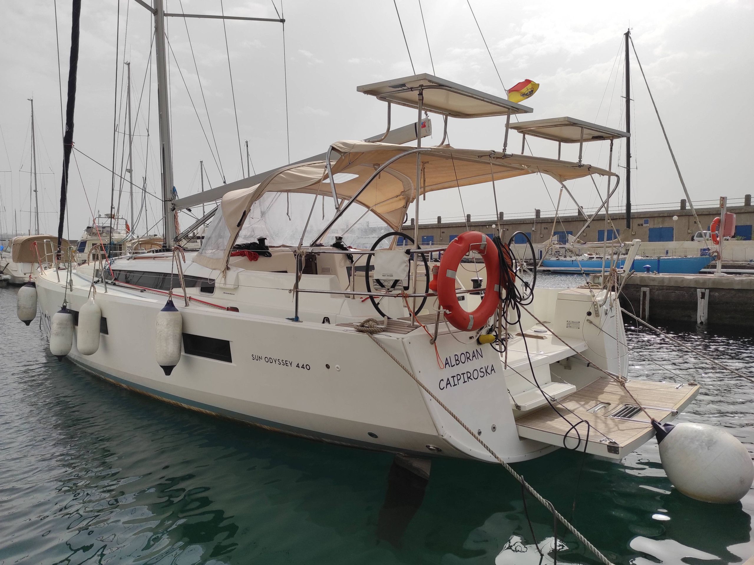 Eine weiße Yacht namens SUN ODYSSEY 440 liegt in einer Marina festgemacht, mit einem Rettungsring und einem Sonnensegel.