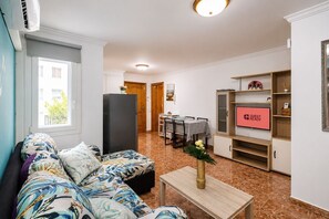 Ein Wohnzimmer mit einer Couch, einem Fernseher, einem Essbereich mit Tisch und Stühlen sowie einer Küche mit Kühlschrank.