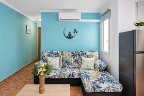 Un salon moderne avec un mur de couleur turquoise, un canapé à motifs floraux, une table basse en bois sur laquelle est posé un vase de fleurs, un réfrigérateur noir et un climatiseur fixé au mur.