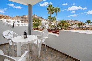 A balcony with white plastic chairs and a table, overlooking a coastal area with palm trees and buildings. - GuestReady - El Cardenal with Pool near Beach (Mogán)