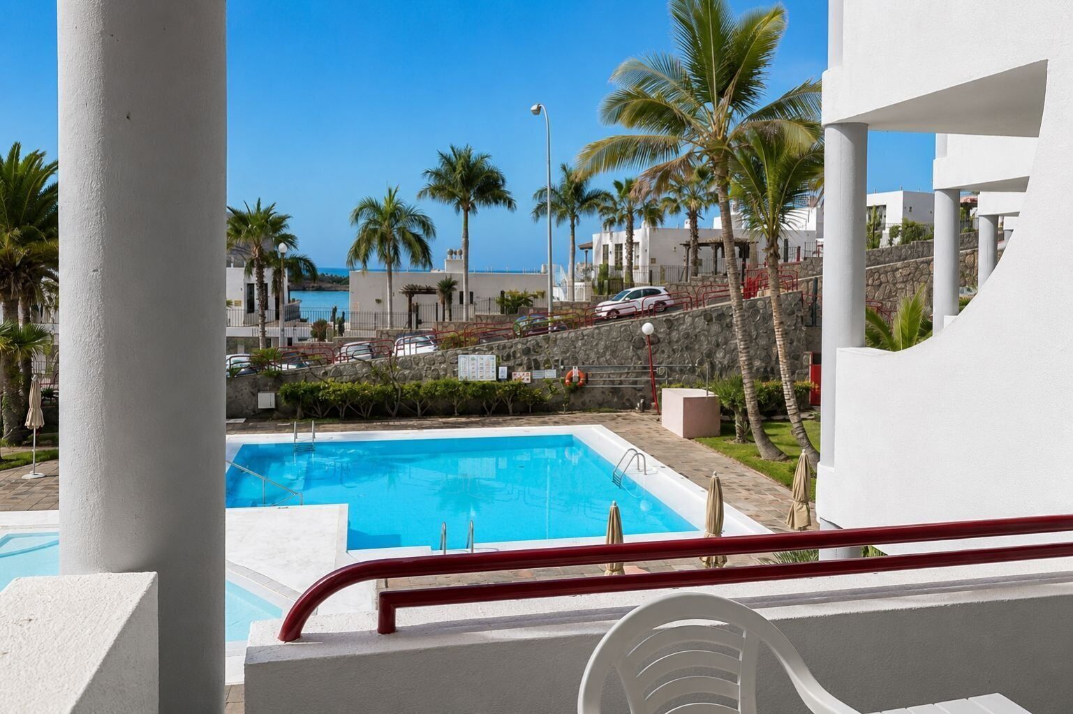 A swimming pool with a red safety railing, palm trees, and buildings in the background.