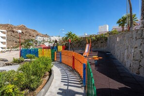Ein Spielplatz mit bunten Spielgeräten und einer Rutsche, umgeben von einer Steinmauer und Pflanzen.