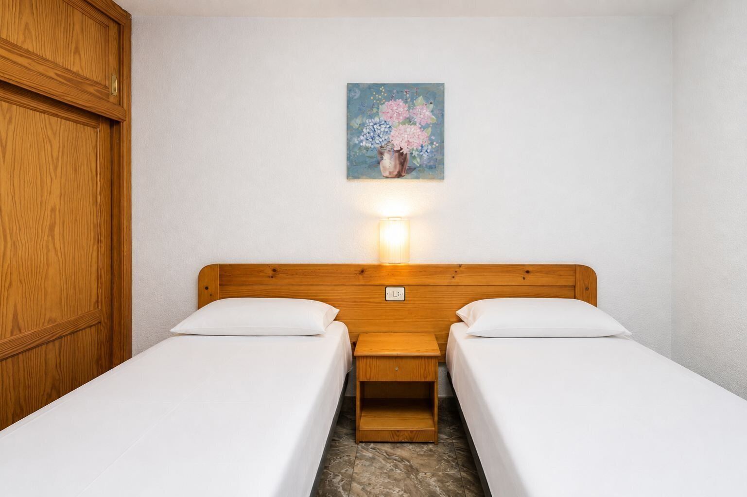 Une chambre avec deux lits, une tête de lit en bois, une table de chevet, une lampe fixée au mur et un tableau représentant des fleurs.