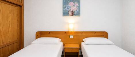 Une chambre avec deux lits, une tête de lit en bois, une table de chevet, une lampe fixée au mur et un tableau représentant des fleurs.