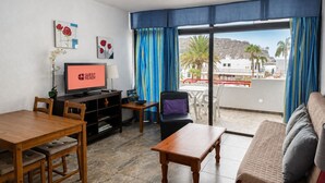 Hotelværelse med spiseplads, sofa, tv der viser 'GUEST READY' og balkon med udsigt til palmer og bygninger.