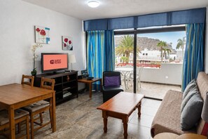 Ein Hotelzimmer mit Essbereich, einer Sitzecke, einem Fernseher, der 'GUEST READY' anzeigt, und einem Balkon mit Blick auf Palmen und Gebäude.