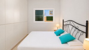 Una cama individual con colchón blanco y dos almohadas color turquesa, cabecera de metal negro, mesita de noche con lámpara y ventana con vista a un edificio.
