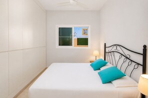 Ein Einzelbett mit weißer Matratze und zwei türkisenen Kissen, ein schwarzes Metall-Kopfstück, ein Nachttisch mit Lampe und ein Fenster mit Blick auf ein Gebäude.